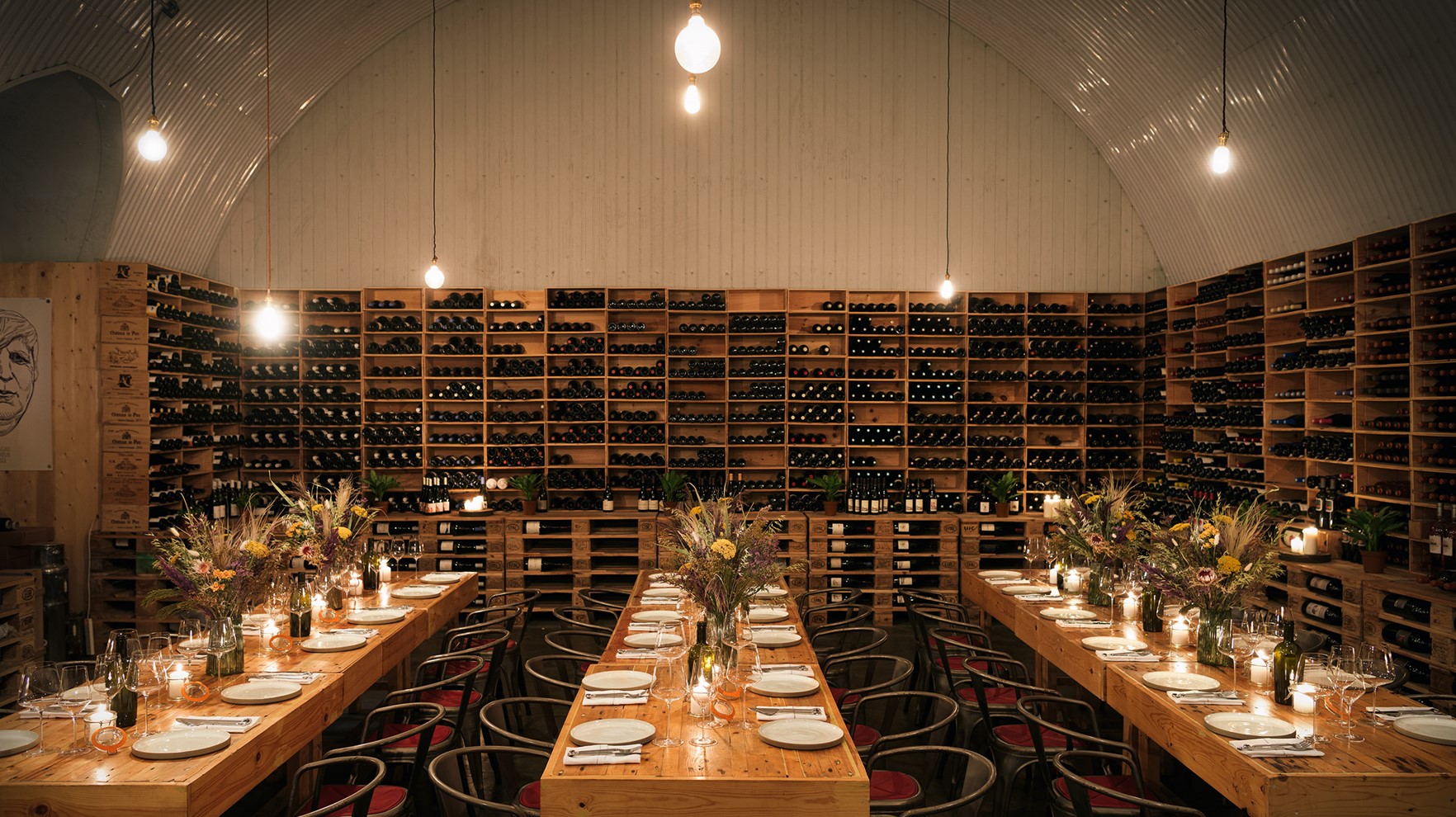 Three rows of tables laid decorated with dinner plates and flowers in a wine warehouse